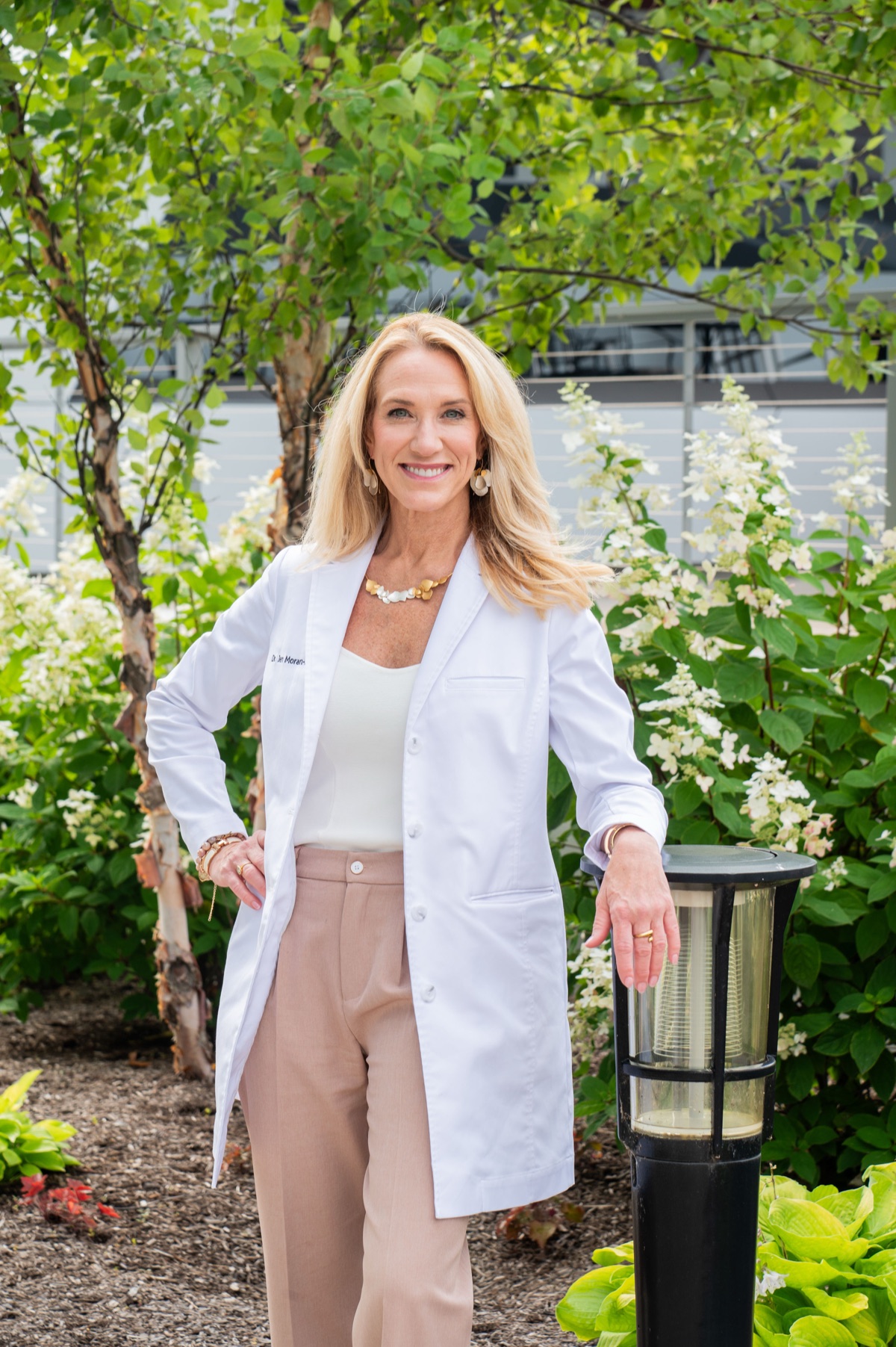 Dr. Jen Moran smiling in her white coat, outdoors among flowering shrubs
